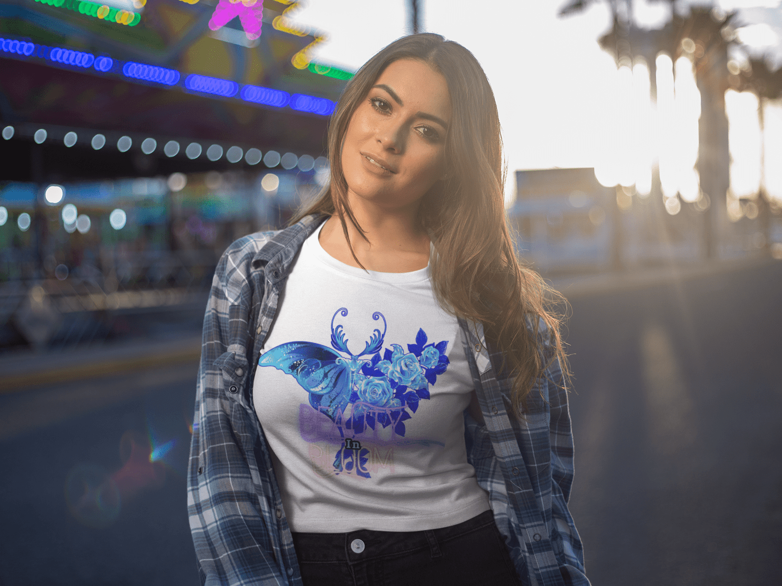A woman posing outdoors during golden hour in the Beauty in Bloom Tee in white, layered with a plaid shirt, highlighting the ethereal butterfly and rose motif.