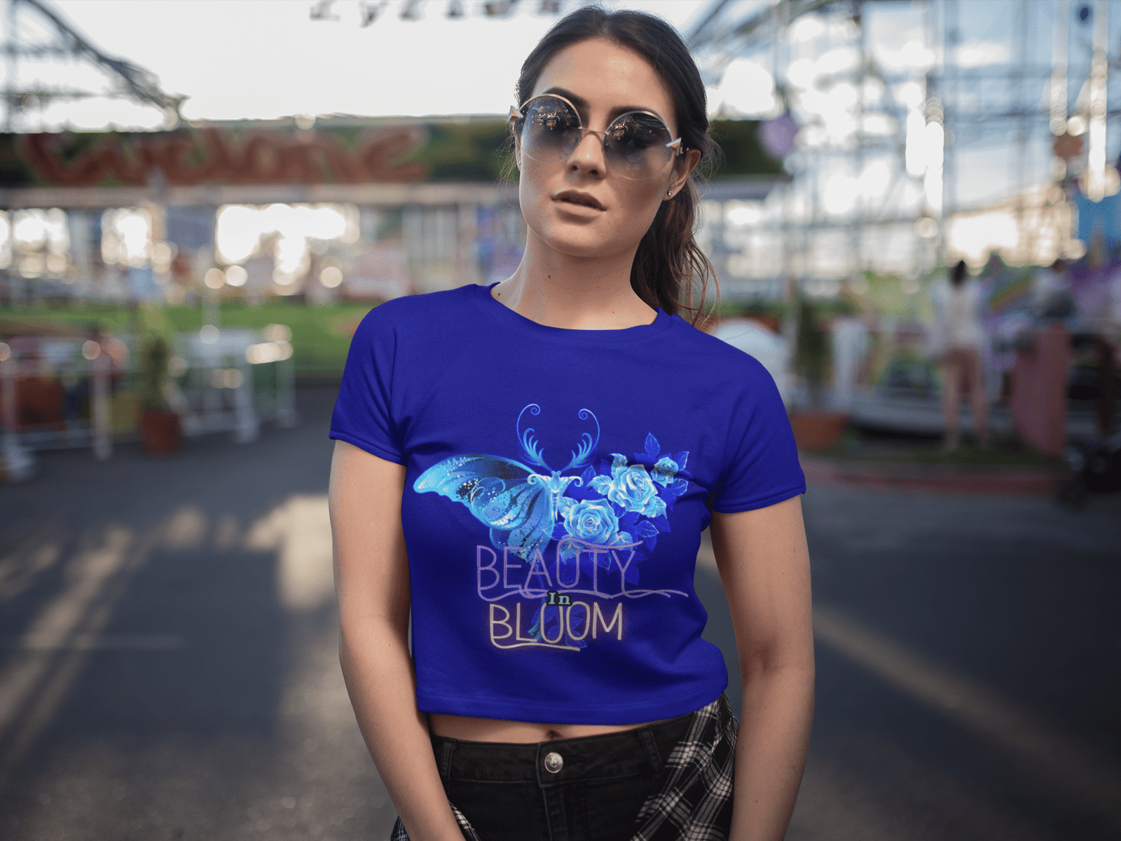 A woman wearing the Beauty in Bloom Tee in royal blue, featuring a vibrant butterfly and rose design, standing confidently at a carnival, embodying a chic and stylish look.