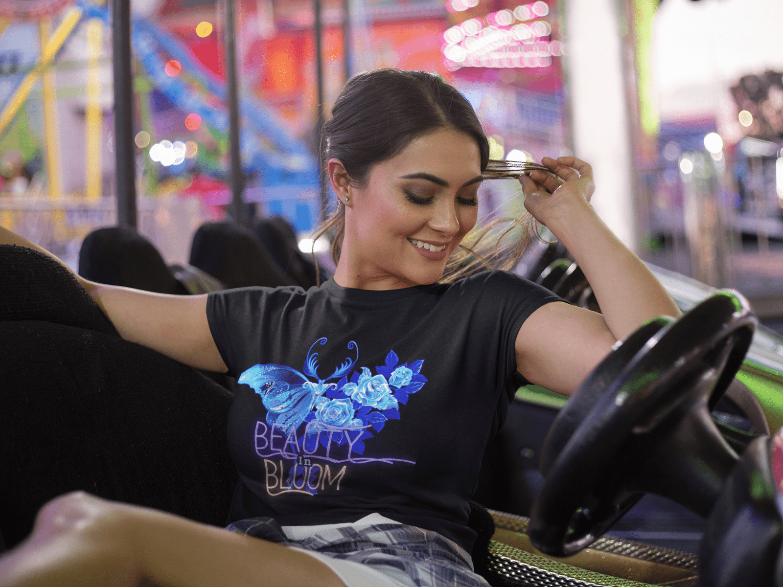 Smiling woman wearing the 'Beauty in Bloom Tee' with a chic rose and butterfly design, enjoying a fun-filled evening at a vibrant amusement park. The black T-shirt showcases a glowing floral and butterfly motif, adding a touch of elegance and style to her casual outfit.