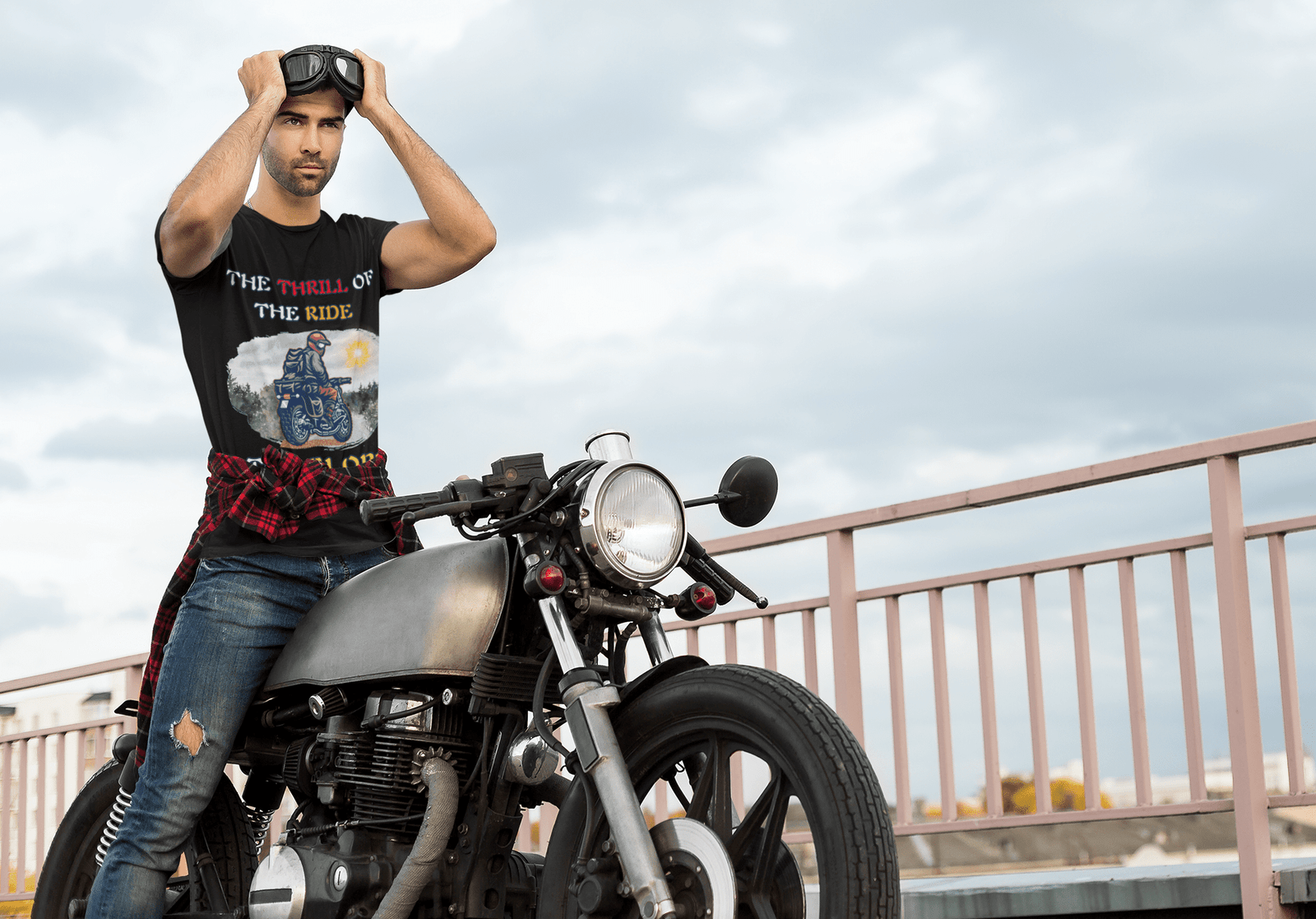 A biker wearing the Thrill Rider tee stands next to a vintage motorcycle on a rooftop, adjusting his goggles under a cloudy sky. The black t-shirt showcases a bold graphic of a fearless motorcyclist on an off-road journey with the words "THE THRILL OF THE RIDE" and "THE GLORY OF THE WIN" in bold typography. Designed for adventure lovers and motorcycle enthusiasts.