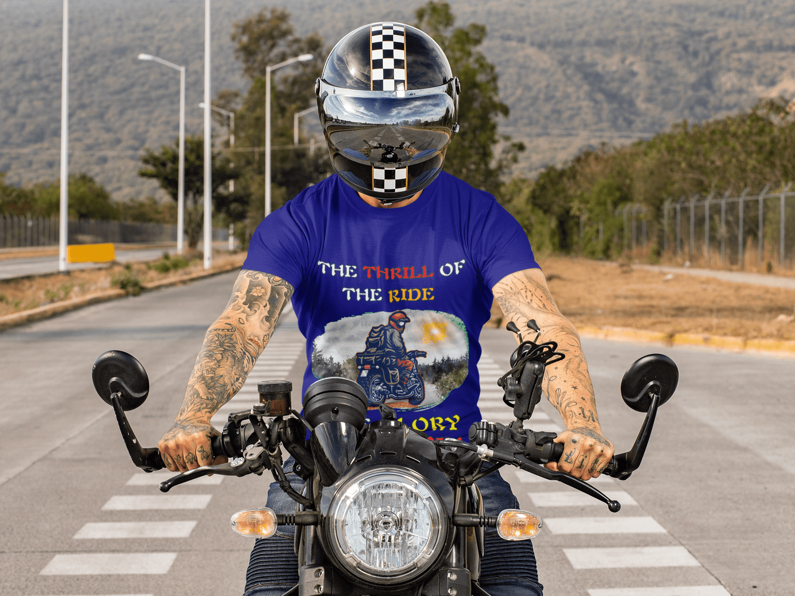 A biker wearing the Thrill Rider tee in navy blue, gripping the handlebars of his motorcycle on an open road. His tattooed arms and checkered racing helmet add to the bold and adventurous look. The t-shirt features a dynamic motorcyclist design with a scenic background and empowering typography, perfect for thrill-seekers and road warriors.