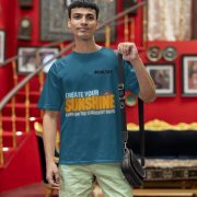 Motivational petrol blue oversized T-shirt with a bold 'CREATE YOUR SUNSHINE EVEN ON THE CLOUDIEST DAYS' design. Vibrant sky-blue, orange/yellow, and white text with a sun icon. DTG-printed on durable 240 GSM cotton fabric. Small size.