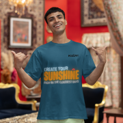 Motivational petrol blue oversized T-shirt with a bold 'CREATE YOUR SUNSHINE EVEN ON THE CLOUDIEST DAYS' design. Vibrant sky-blue, orange/yellow, and white text with a sun icon. DTG-printed on durable 240 GSM cotton fabric. Small size.