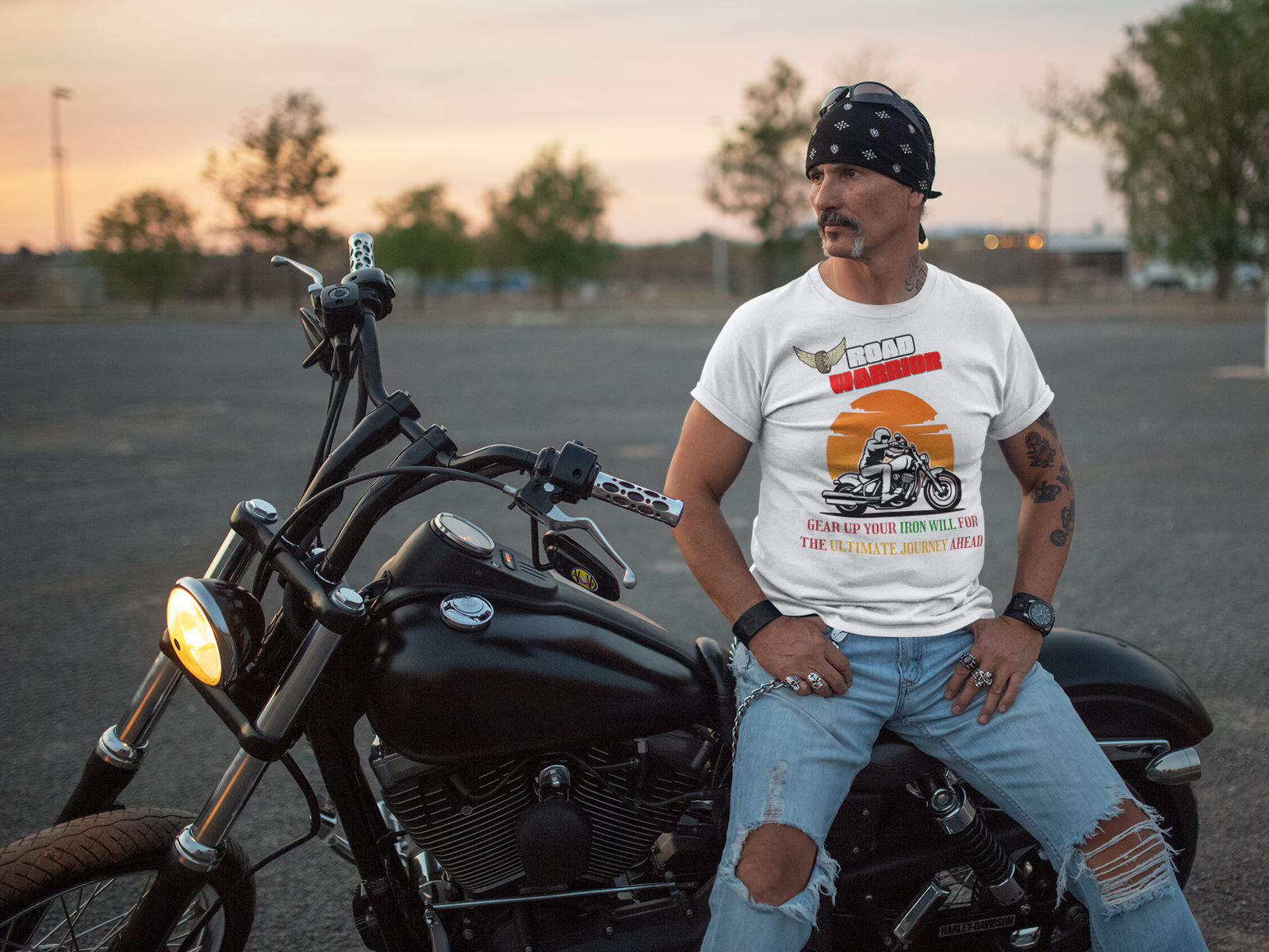 A rugged biker wearing the white Road Warrior tee, sitting on a black cruiser motorcycle at sunset. The tee features a bold motorcyclist design against a blazing orange sunset, with "ROAD WARRIOR" in red and white text, and a motivational phrase in vibrant colors. Perfect for adventure seekers and motorcycle enthusiasts.