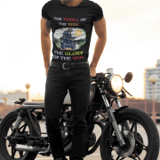 A stylish biker wearing the Thrill Rider Tee by PodLyft, standing confidently next to a classic black motorcycle. The black t-shirt features a bold design of a fearless motorcyclist riding through a scenic outdoor backdrop with a bright sun, green landscapes, and a winding road. The typography highlights "THE THRILL OF THE RIDE" and "THE GLORY OF THE WIN" in bold mixed colors, capturing the essence of adventure and freedom. A perfect tee for motorcycle enthusiasts, road warriors, and thrill-seekers who embrace the open road with passion and style.