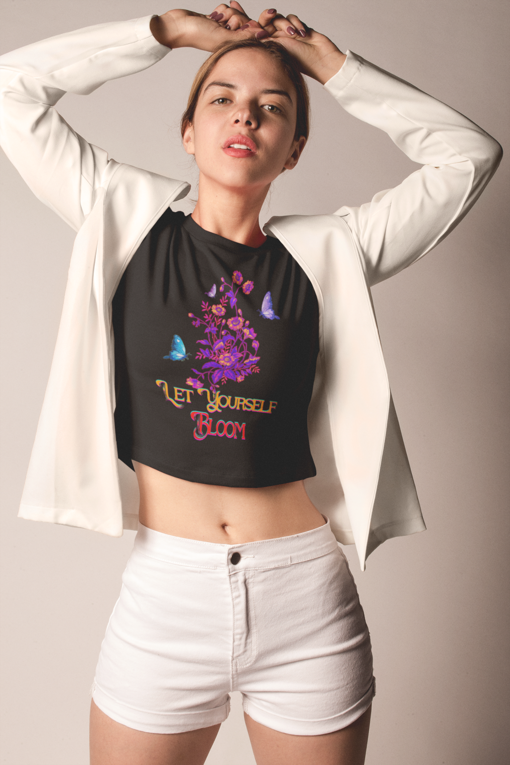 Confident woman wearing a stylish black crop top with a bold 'Bloom Yourself Tee' floral and butterfly design. She pairs it with a chic white blazer and shorts, creating a modern and fashionable look. Available at PodLyft.