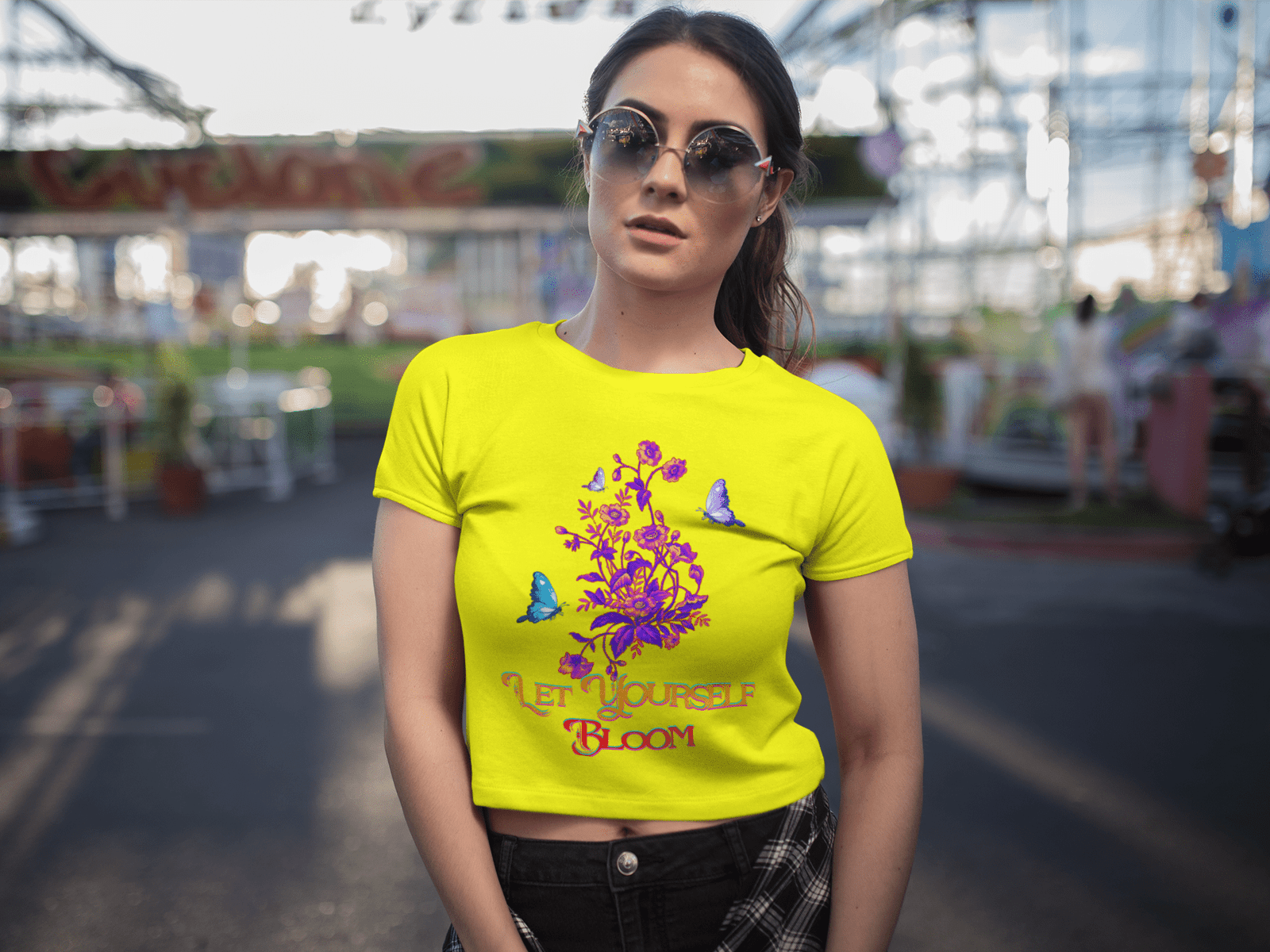 Stylish woman wearing a vibrant golden yellow Bloom Yourself Tee featuring a bold 'Let Yourself Bloom' floral and butterfly design. She pairs it with trendy sunglasses and black jeans, standing confidently in a lively amusement park setting. Available at PodLyft.