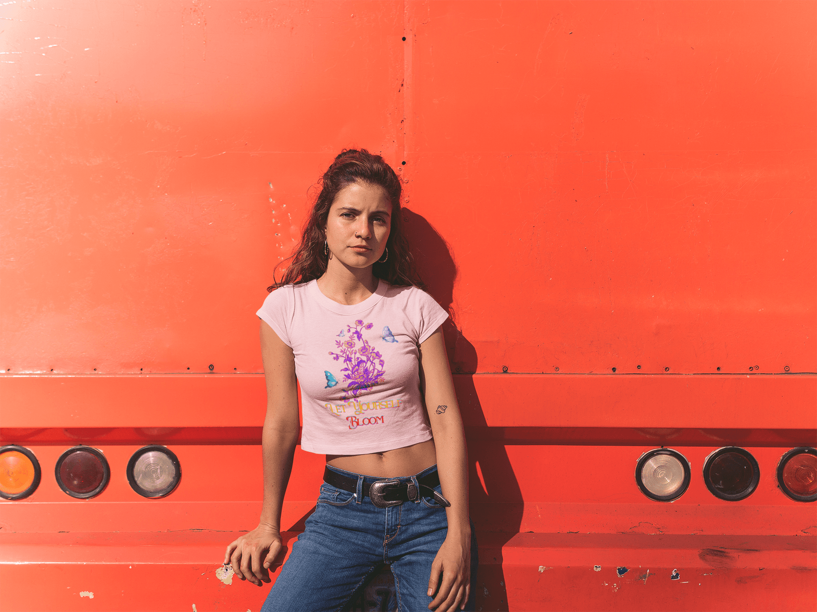 Confident young woman wearing a soft pink Bloom Yourself Tee featuring the 'Let Yourself Bloom' floral and butterfly design. She pairs it with blue jeans and a black belt, leaning against a bold orange backdrop. Available at PodLyft.
