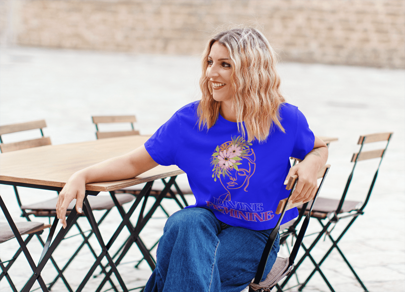 Smiling woman wearing a vibrant blue Bloom Yourself Tee featuring a minimalist floral face design with the words 'Divine Feminine.' She is seated at an outdoor café, exuding confidence and elegance. Available at PodLyft.