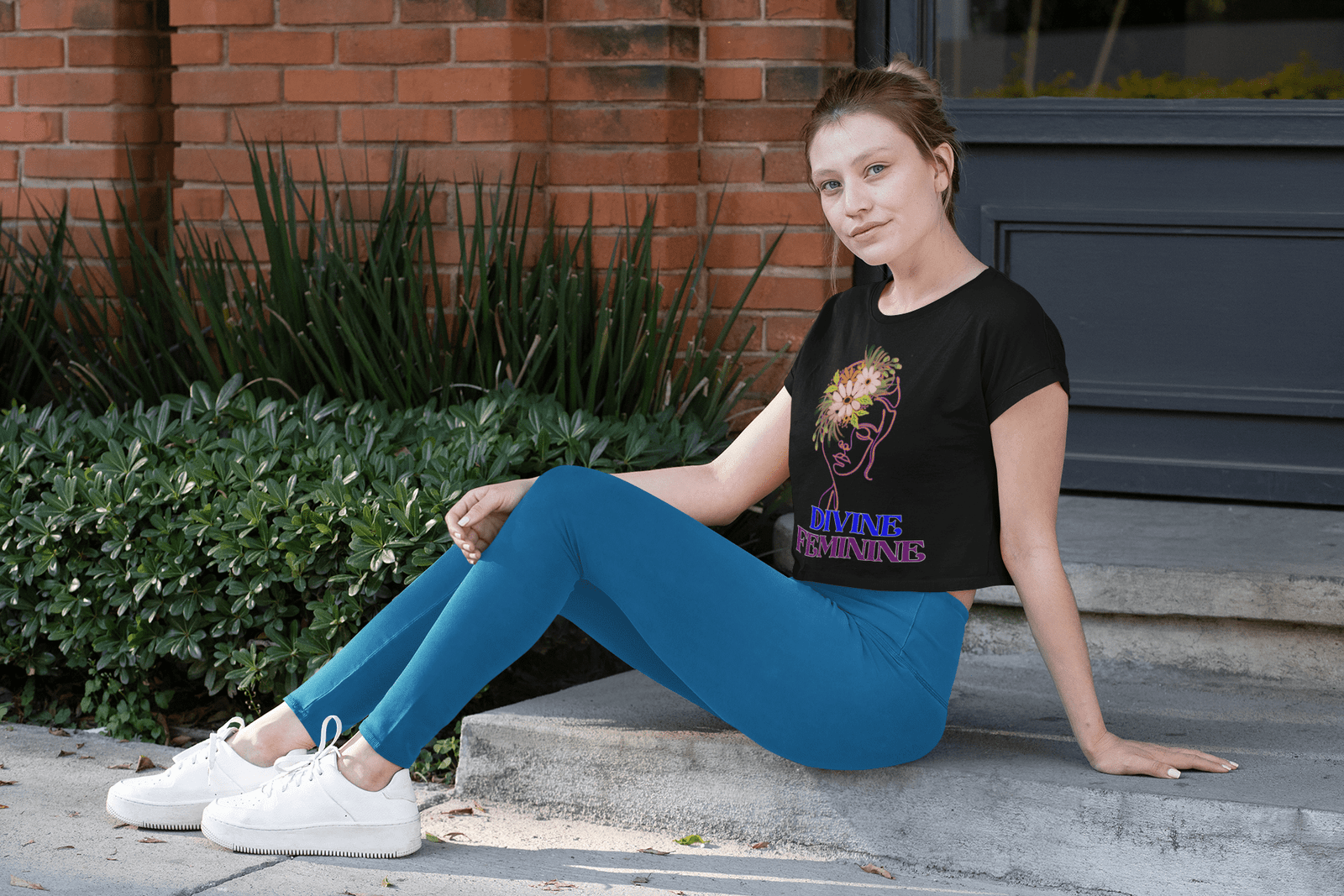 Woman wearing a black Divine Feminine T-shirt with a floral line-art design, sitting casually on outdoor steps. A stylish and empowering nature-inspired tee, available at PodLyft.