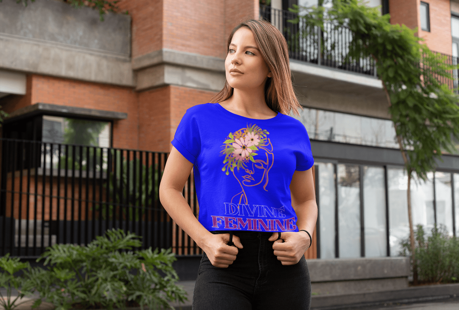 Woman wearing a bold blue Divine Feminine T-shirt featuring an elegant floral line-art design, standing confidently in an urban setting. A stylish and empowering statement tee, available at PodLyft.