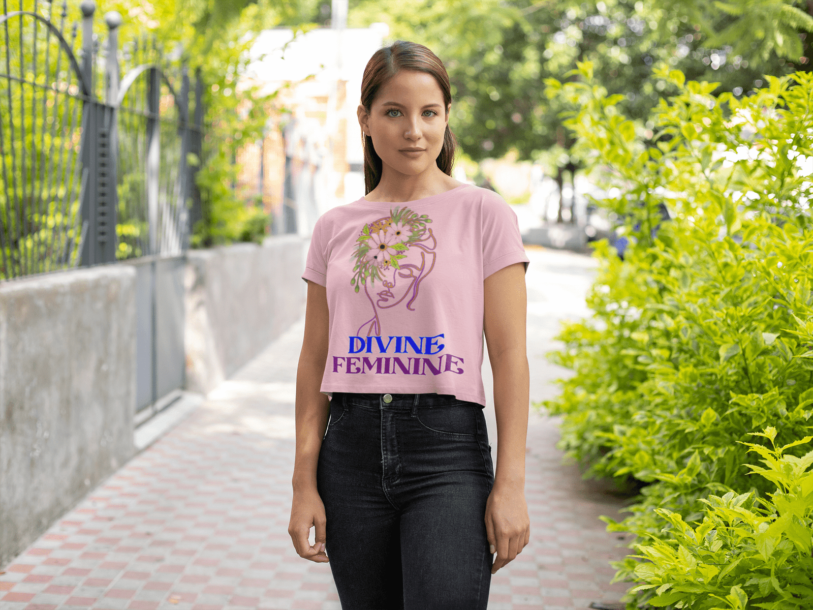 Woman wearing a soft pink Divine Feminine T-shirt featuring a delicate floral line-art design, walking along a stylish outdoor pathway. A chic and empowering fashion piece, available at PodLyft.