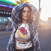 A stylish young woman poses confidently at an outdoor fairground, wearing a white Elegant Floral Crown T-shirt featuring an elegant floral crown design with the words 'Crowned by Nature.' She pairs the T-shirt with a plaid shirt and high-waisted black jeans, illuminated by warm sunset lighting. The vibrant background adds a dynamic urban aesthetic to the scene.