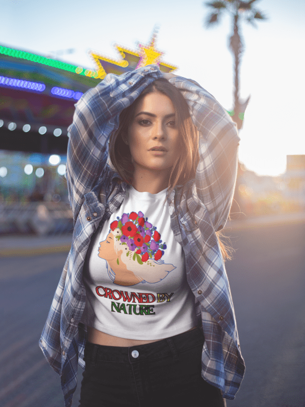 A stylish young woman poses confidently at an outdoor fairground, wearing a white Elegant Floral Crown T-shirt featuring an elegant floral crown design with the words 'Crowned by Nature.' She pairs the T-shirt with a plaid shirt and high-waisted black jeans, illuminated by warm sunset lighting. The vibrant background adds a dynamic urban aesthetic to the scene.