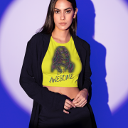A stylish model wearing the I'm Awesome crop top in bold golden yellow, featuring a vibrant neon-style artistic design of a confident woman and striking typography. The outfit is paired with a black blazer and pants, set against a dramatic purple and white spotlight background, creating a powerful and fashionable statement.
