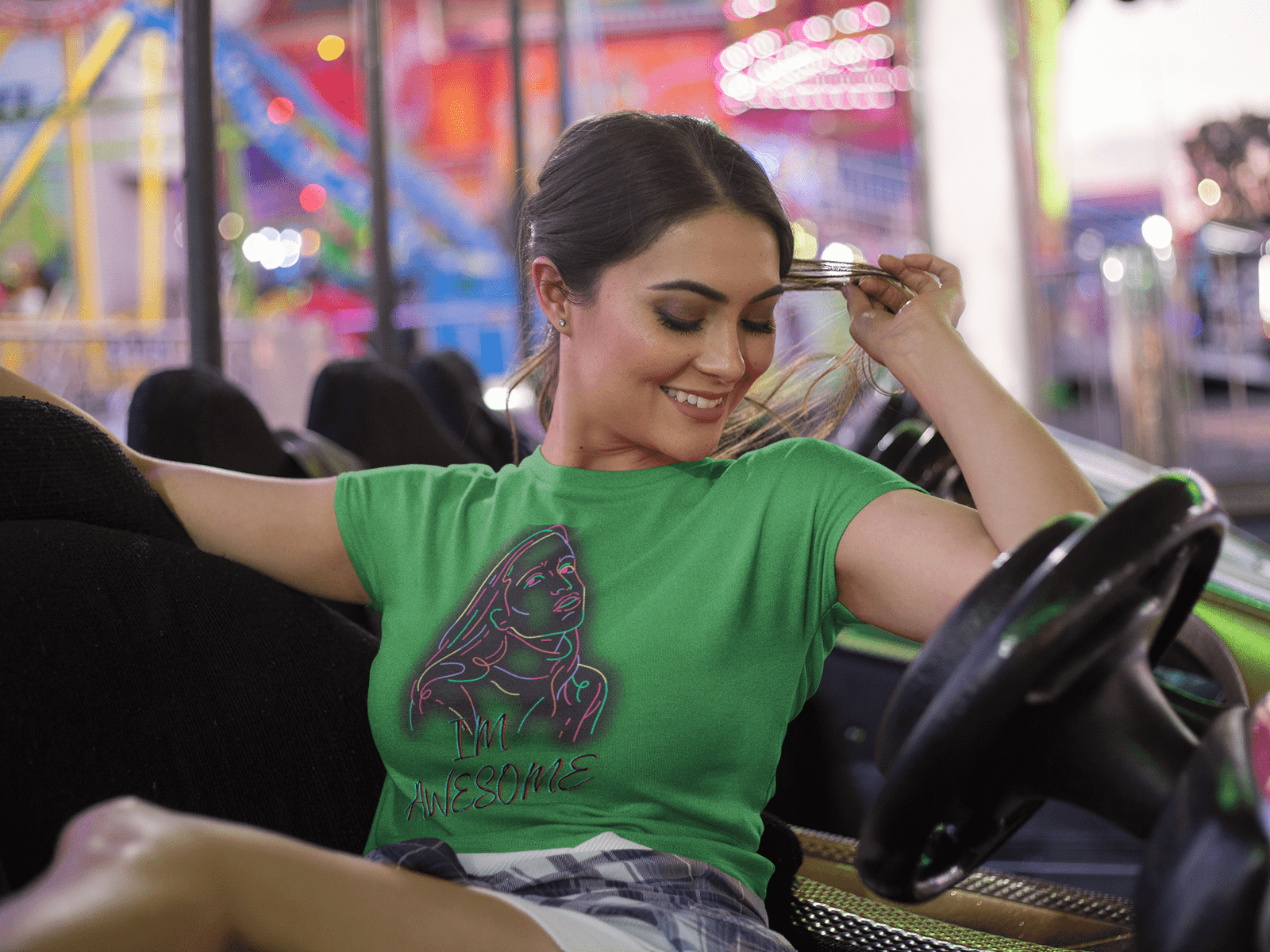 A cheerful young woman wearing the I'm Awesome crop top in vibrant green, featuring a neon-style artistic portrait and bold typography. She is relaxing in a bumper car at a lively amusement park, smiling joyfully with bright carnival lights in the background, exuding confidence and fun-loving energy.