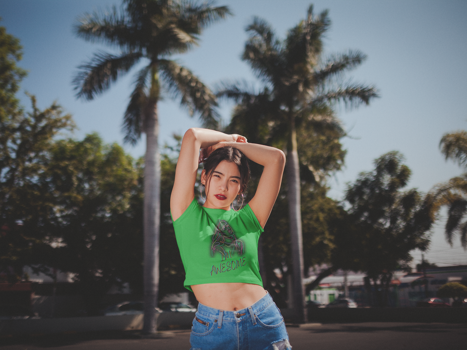 A confident young woman poses stylishly in the I'm Awesome crop top in bold green, featuring a neon-style artistic portrait and empowering typography. She stands outdoors in a tropical setting with palm trees in the background, exuding a carefree and self-assured vibe. The high-waisted denim shorts complement the trendy, modern aesthetic of the outfit.