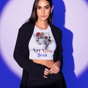 A stylish white Let Love Soar Tee featuring an elegant design of two doves carrying roses with the text "Let Love Soar" in a mix of red, black, and blue fonts. Perfect for expressing love, freedom, and passion. Modeled with a chic black blazer for a bold yet romantic look. High-quality apparel by PodLyft.
