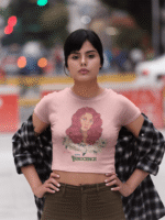 Model in a baby pink 'Beauty with Innocence' crop top by PodLyft, featuring a red-haired character with floral accents and pink-green text, paired with light blue high-waisted pants, standing on a beach at dusk with mountains in the background.