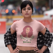 Model in a baby pink 'Beauty with Innocence' crop top by PodLyft, featuring a red-haired character with floral accents and pink-green text, paired with light blue high-waisted pants, standing on a beach at dusk with mountains in the background.