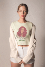 Model wearing a beige 'Beauty with Innocence' crop top by PodLyft, highlighting a red-haired character with white floral details and pink-green text, styled with light blue pants, against a soft sunset with a distant ocean and mountainous horizon.