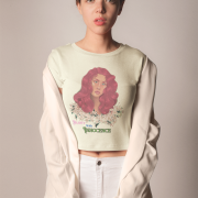 Model wearing a beige 'Beauty with Innocence' crop top by PodLyft, highlighting a red-haired character with white floral details and pink-green text, styled with light blue pants, against a soft sunset with a distant ocean and mountainous horizon.