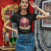 Model wearing a black 'Beauty with Innocence' crop top by PodLyft, featuring a red-haired character with white floral accents and pink-green text, paired with light blue high-waisted pants, standing against a serene sunset with mountains and water in the background.