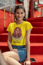 Model in a golden yellow 'Beauty with Innocence' crop top by PodLyft, displaying a red-haired character with floral details and pink-green text, styled with light blue pants, set against a sunset backdrop with distant mountains and a calm ocean.