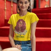 Model in a golden yellow 'Beauty with Innocence' crop top by PodLyft, displaying a red-haired character with floral details and pink-green text, styled with light blue pants, set against a sunset backdrop with distant mountains and a calm ocean.