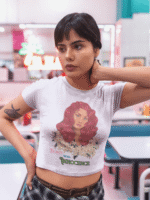 Model in a lavender 'Beauty with Innocence' crop top by PodLyft, displaying a red-haired character with floral elements and pink-green text, paired with light blue trousers, set against a sunset backdrop with mountains and a serene water view.