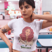 Model in a lavender 'Beauty with Innocence' crop top by PodLyft, displaying a red-haired character with floral elements and pink-green text, paired with light blue trousers, set against a sunset backdrop with mountains and a serene water view.