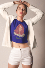 Model wearing a navy blue 'Beauty with Innocence' crop top by PodLyft, featuring a red-haired character with white floral accents and pink-green text, paired with light blue high-waisted pants, standing against a serene sunset with mountains and water in the background.