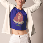 Model wearing a navy blue 'Beauty with Innocence' crop top by PodLyft, featuring a red-haired character with white floral accents and pink-green text, paired with light blue high-waisted pants, standing against a serene sunset with mountains and water in the background.