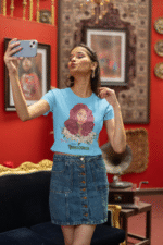 Model showcasing a sky blue 'Beauty with Innocence' crop top by PodLyft, with a red-haired character design, white flowers, and pink-green text, wearing light blue trousers, posed against a tranquil sunset with mountains and water in the distance.