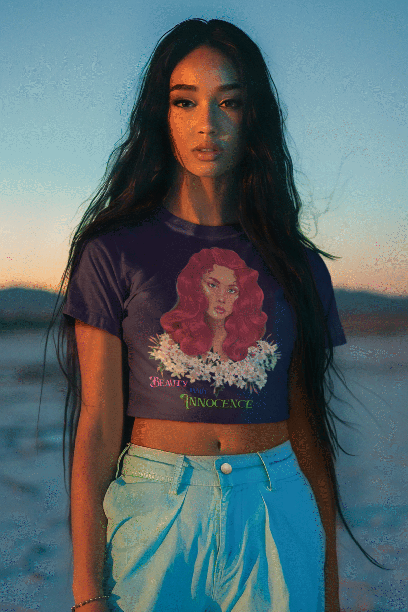 Model wearing a navy blue Beauty with Innocence crop top by PodLyft, featuring a red-haired character with floral accents and text in pink and green, paired with light blue high-waisted pants, standing against a serene sunset backdrop with mountains and water in the distance.