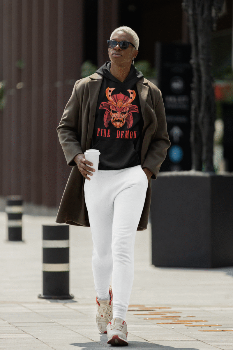 Men’s Fire Demon Samurai Hoodie by PodLyft in black, on a mannequin, featuring a bold red and orange samurai helmet design with a demonic face and 'FIRE DEMON' text, displayed in a modern store with blurred clothing racks in the background.