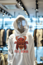 Men’s Fire Demon Samurai Hoodie by PodLyft on a mannequin, white fabric with a bold red and orange samurai helmet design featuring a demonic face and 'FIRE DEMON' text, displayed in a modern store with blurred clothing racks in the background.
