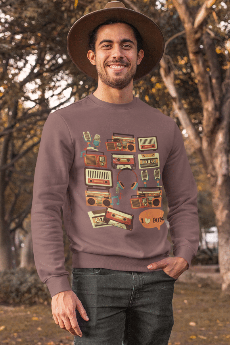 Man wearing a brown 90s retro sweatshirt featuring vintage cassette tapes, boomboxes, radios, and music icons, standing outdoors with a cheerful smile and autumn trees in the background — PodLyft retro sweatshirt for men.