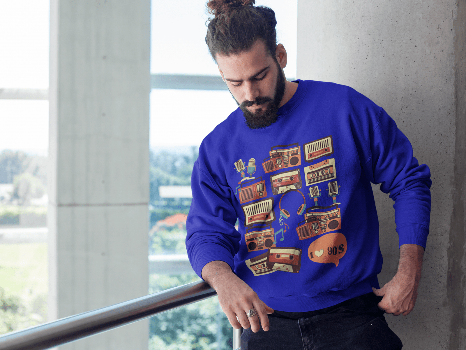 Bearded man in a vibrant royal blue 90s retro sweatshirt featuring colorful nostalgic music graphics, standing indoors near a window with a city view.