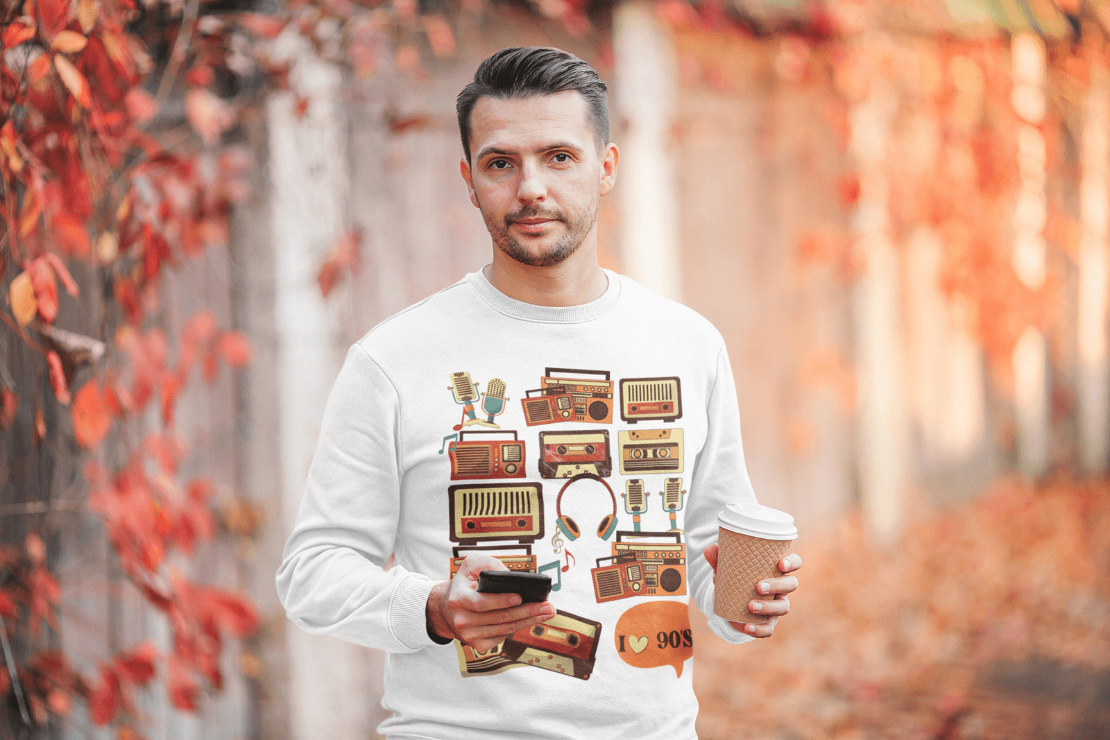 Man wearing a white 90s retro sweatshirt with vintage cassette and radio graphics, holding a coffee cup and smartphone, standing outdoors with red autumn leaves in the background.