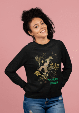 Smiling woman wearing the Nature in My Hands Sweatshirt, a black eco-friendly garment featuring a symbolic design of a hand intertwined with green and gold botanical elements, mystical stars, and the phrase "NATURE IN MY HANDS" in bold green text; posed against a soft pink background.