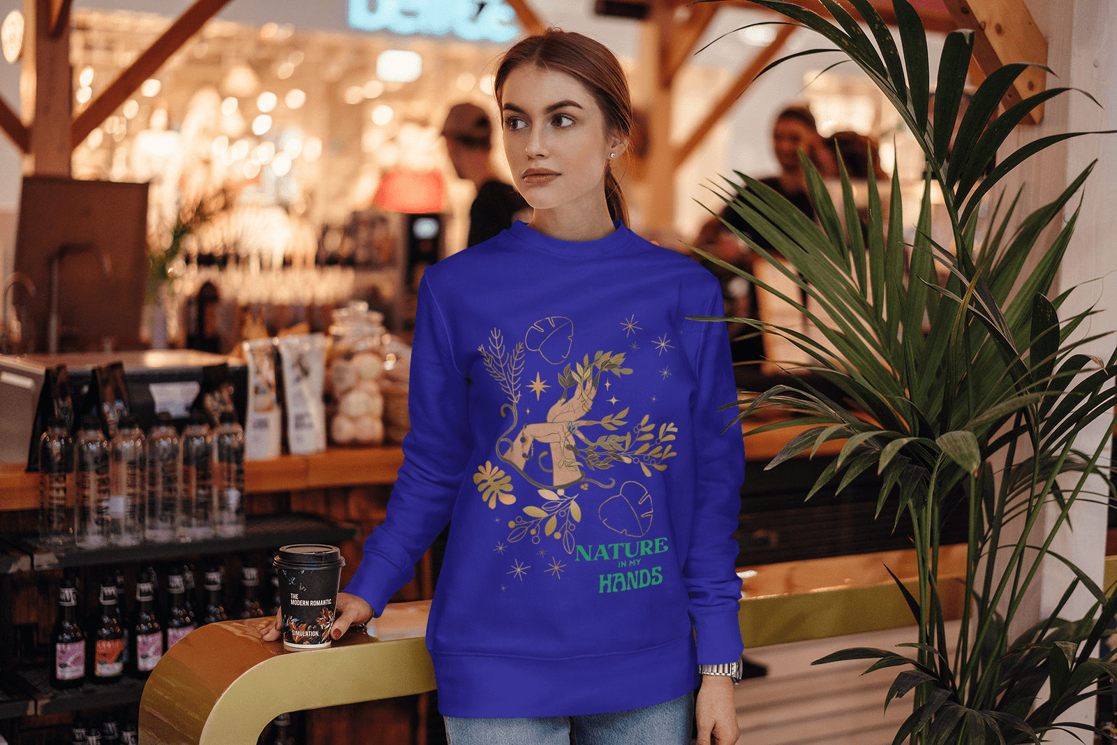 Young woman in the Nature in My Hands Sweatshirt holding a coffee cup inside a cozy café, surrounded by warm lighting and greenery.