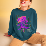 Woman wearing a teal sweatshirt with a vibrant neon floral print and the phrase 'BLOOM WHERE YOU'RE PLANTED'; styled with tan joggers, chunky gold jewelry, and oversized orange sunglasses, sitting confidently against a warm beige background. PodLyft Vibrant Floral Sweatshirt for Women.