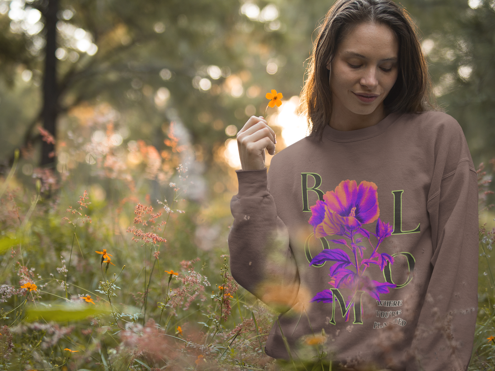 A serene woman stands in a lush field of wildflowers, holding a small orange flower while wearing a warm brown Vibrant Floral Sweatshirt. The sweatshirt displays a vivid floral design in purple and pink with the word “BLOOM” and the phrase “Where You're Planted” integrated into the design.