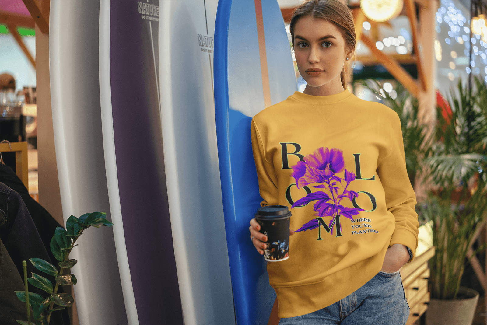 A stylish woman stands indoors holding a coffee cup, wearing a mustard yellow Vibrant Floral Sweatshirt featuring a vibrant purple-pink flower and green typography spelling “BLOOM” with the quote “Where You're Planted.” She stands against a backdrop of colorful surfboards and decorative plants.