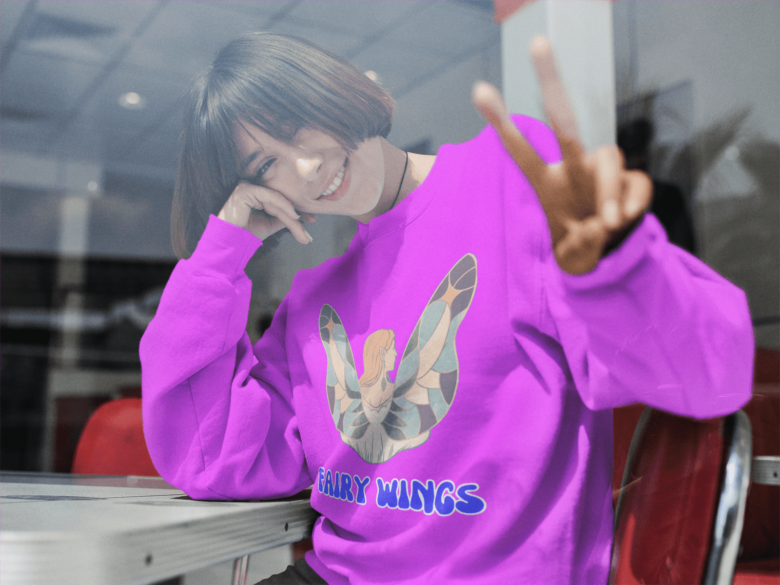 Smiling woman in a vibrant pink Fairy Wings Hoodie showing a peace sign at a café table, showcasing the fantasy wing artwork.