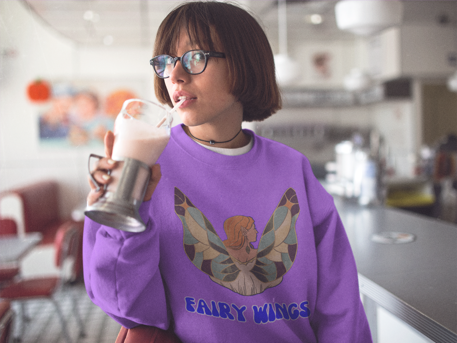 Woman wearing a purple Fairy Wings Hoodie featuring a colorful fairy design, sipping a milkshake in a retro diner.