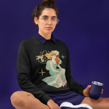 Woman wearing the Women’s Nature Muse Sweatshirt from PodLyft – featuring a vintage-style Art Nouveau design with a red-haired muse, flowers, and a hummingbird, sitting casually against a deep violet background.
