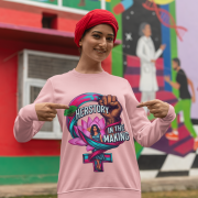 A smiling woman wearing a red turban and a baby pink PodLyft Feminist Power Sweatshirt, featuring a bold graphic with a female symbol, a lotus flower, a raised fist, and the text "HERSTORY IN THE MAKING," pointing to the design in front of a colorful mural.