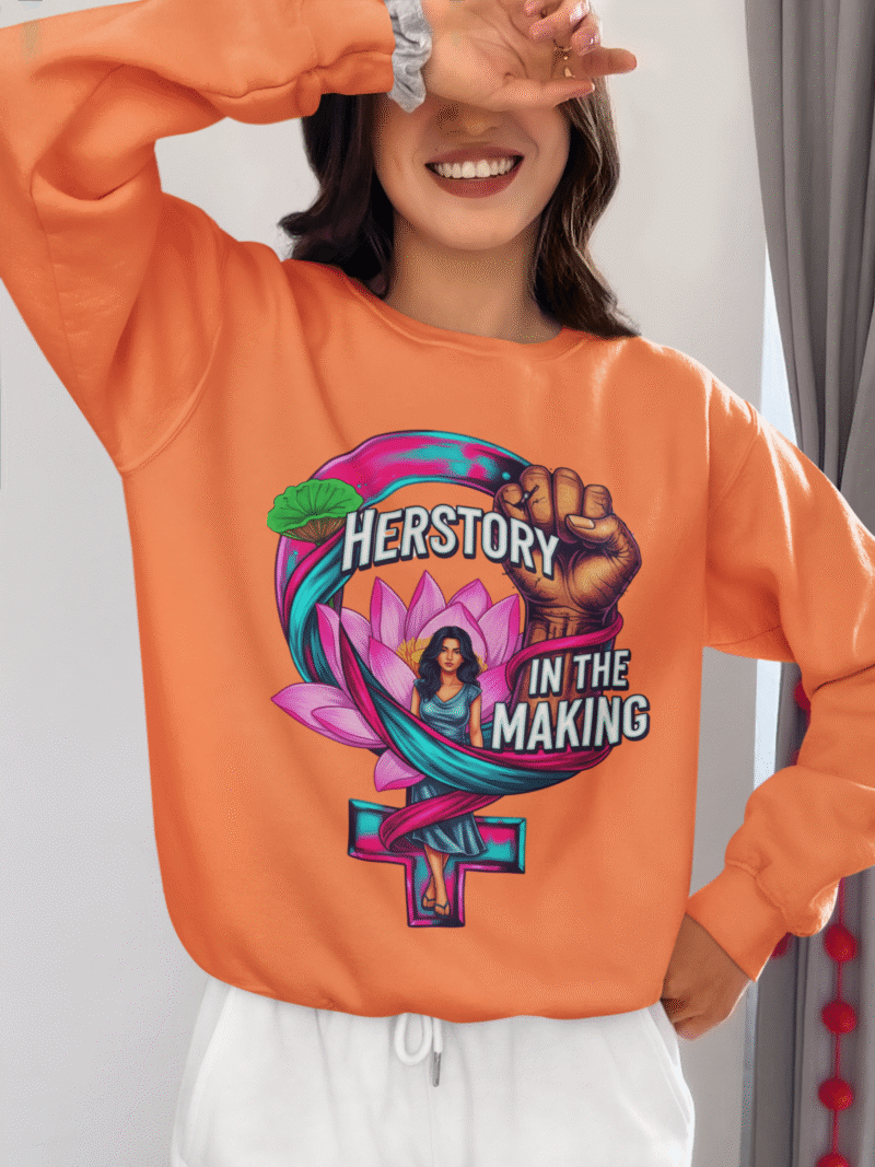 A woman smiling, covering her face with one hand, wearing a coral PodLyft Feminist Power Sweatshirt, featuring a bold graphic with a female symbol, a lotus flower, a raised fist, and the text "HERSTORY IN THE MAKING.